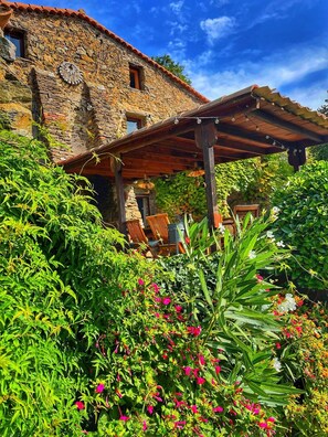 Un bâtiment en pierre agrémenté d’une pergola en bois, entouré d’une végétation luxuriante et de plantes en fleurs.