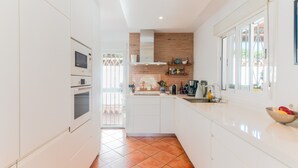Une cuisine moderne avec des meubles blancs, un mur de fond en briques et une fenêtre donnant sur un balcon.