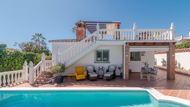Un espace aménagé au bord de la piscine, avec une balustrade blanche, un escalier menant à un balcon et du mobilier d’extérieur.