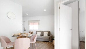 Un salon moderne avec une table ronde en verre, deux fauteuils roses, un canapé gris et une porte blanche.