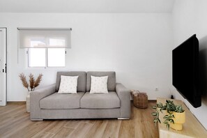 Una sala de estar moderna con un sofá gris, un televisor de pantalla plana, una ventana con persiana blanca y piso de madera.