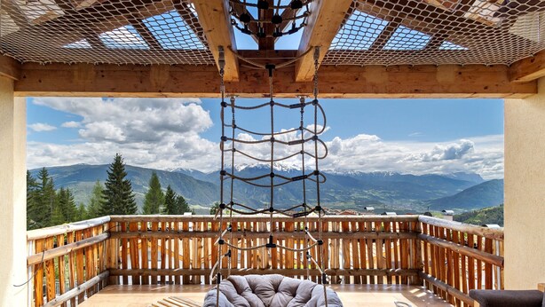 A wooden deck with a hanging swing chair, a cushioned seating area, and a view of mountains.