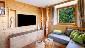 A wooden interior with a flat-screen TV, wooden cabinets, a sofa with cushions, and a window with a forest view.