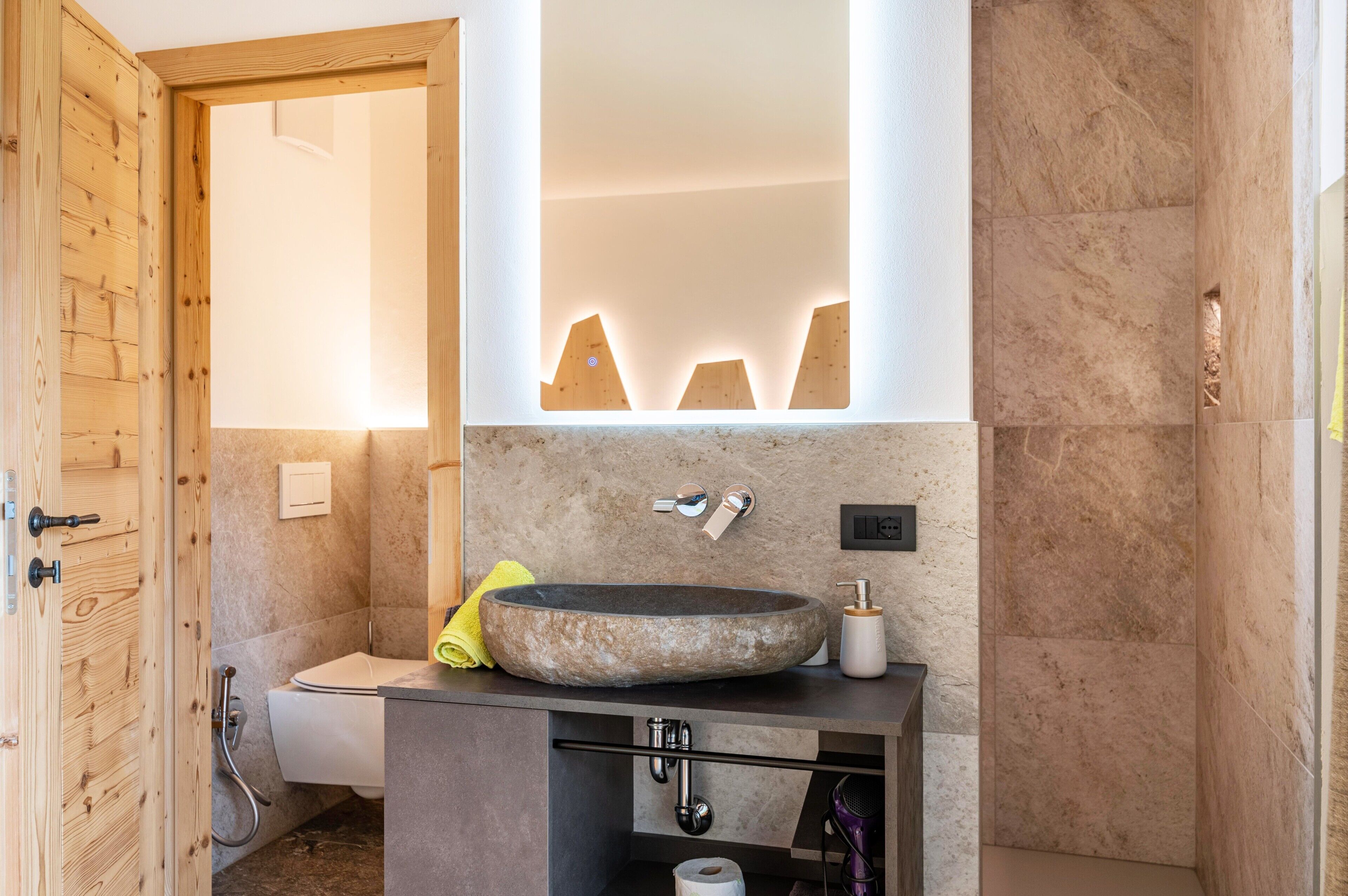 A bathroom with a stone sink, wooden door, and a mirror with lighted edges.