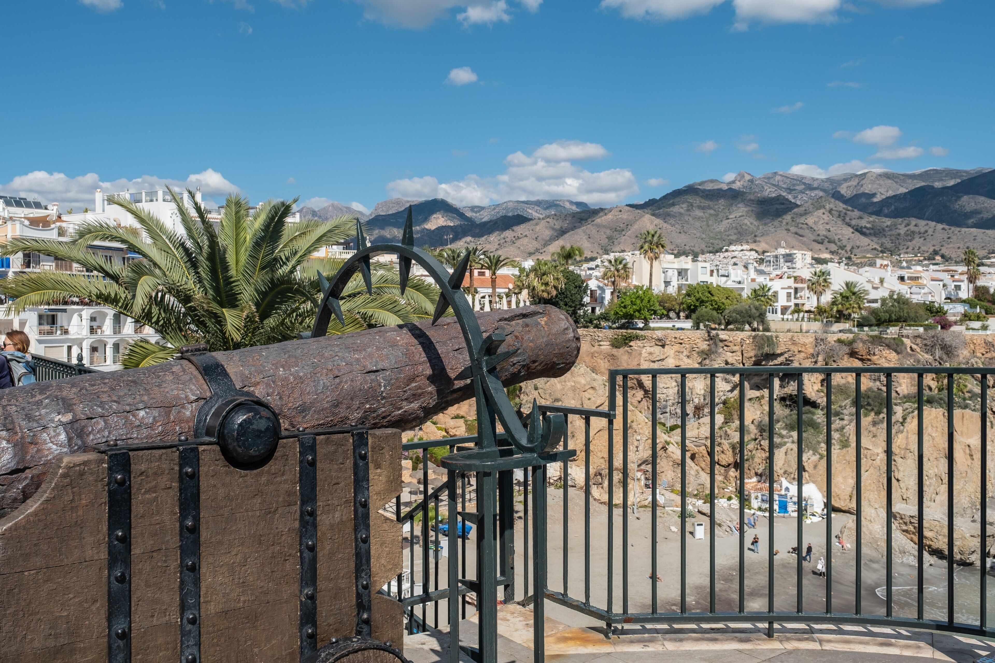 A coastal view with a large cannon, a metal gate, and buildings in the background.