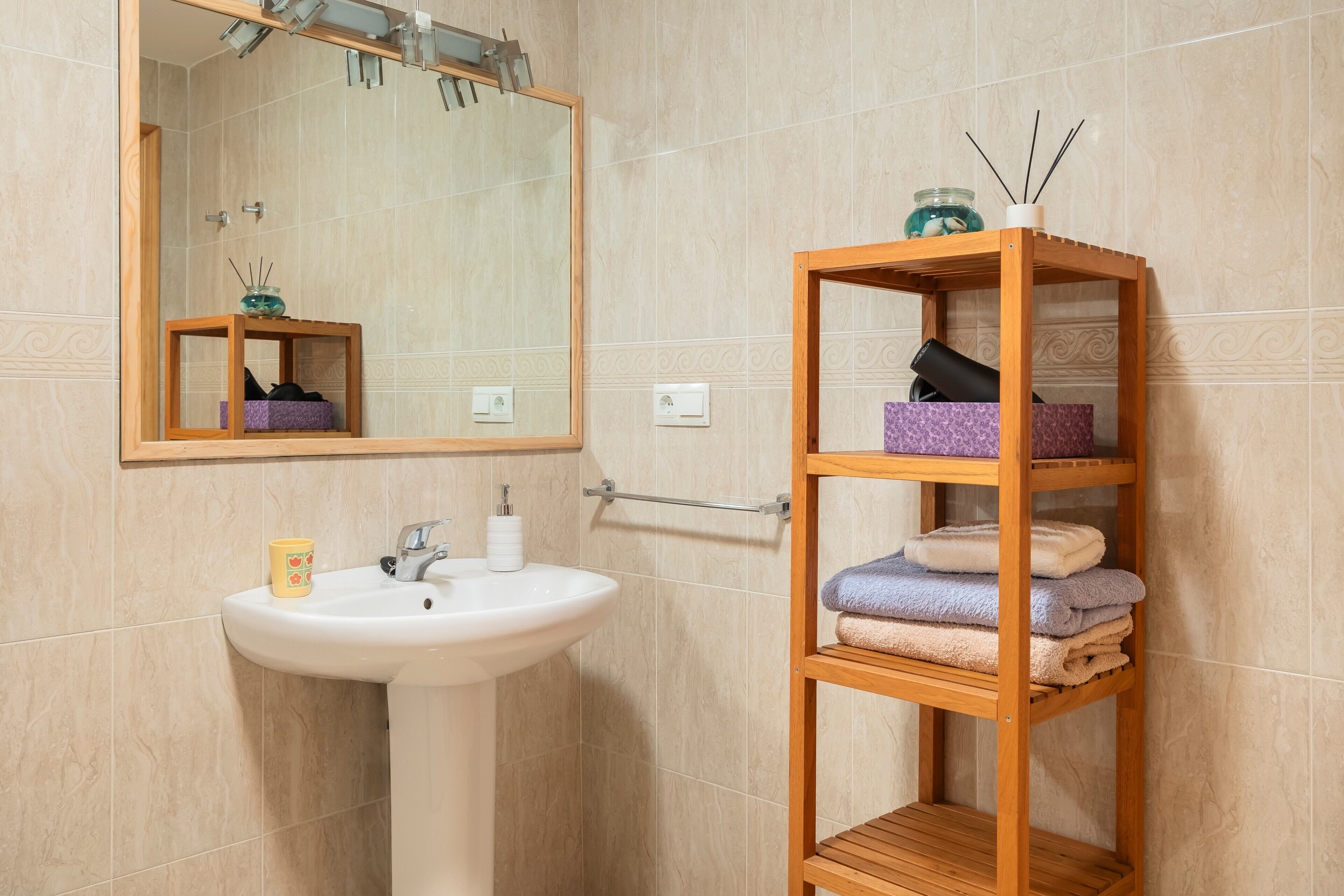 Ein Badezimmer mit Waschbecken, Spiegel, Holzablage mit Handtüchern und einer Dekojar.