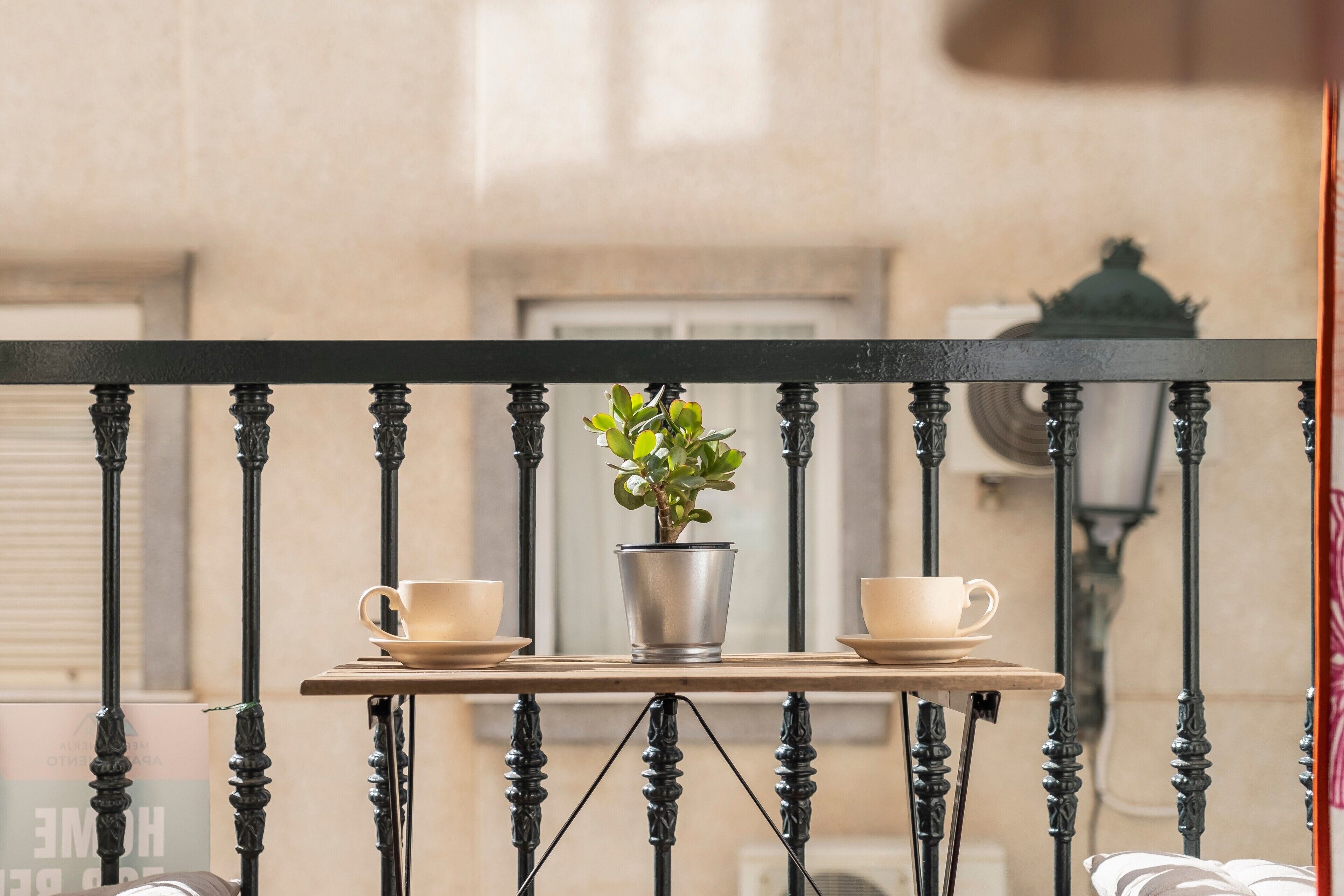 Un balcón con barandilla de metal, una mesa pequeña con una planta en maceta y dos tazas.