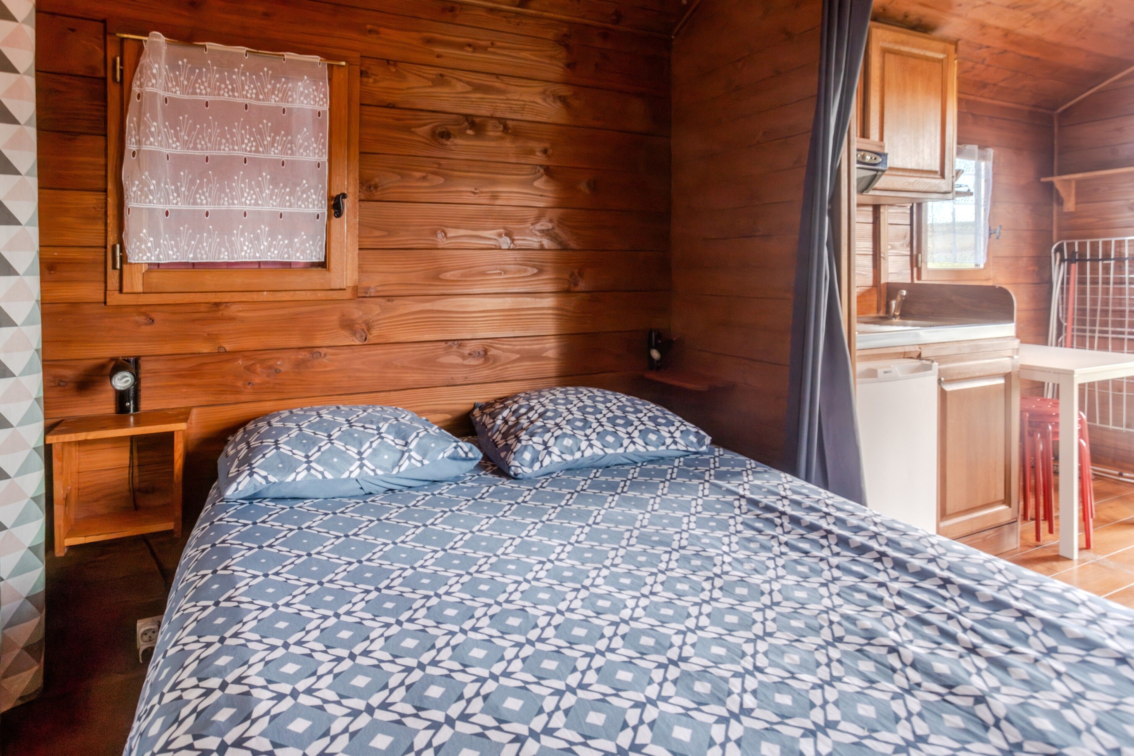 Interno di una baita in legno con un letto, un piccolo comodino, una finestra con tenda e un angolo cottura.