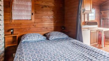 Interno di una baita in legno con un letto, un piccolo comodino, una finestra con tenda e un angolo cottura.