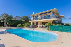 Une maison à deux étages avec une piscine, du mobilier d’extérieur et une clôture verte.
