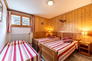 Une chambre avec deux lits simples, une tête de lit en bois, une fenêtre avec des rideaux, un radiateur et une lampe fixée au mur.