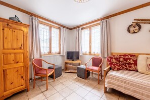 Une pièce comprenant un meuble en bois, deux chaises, un téléviseur posé sur un meuble en bois, un canapé avec des coussins à motifs et une fenêtre avec des rideaux.