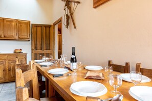Une table de salle à manger en bois, mise avec des assiettes, des verres et une bouteille de vin, entourée de chaises en bois dans une cuisine dont les meubles sont également en bois.