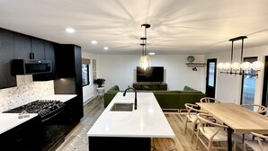 A modern kitchen with a central island, a dining area with a wooden table and chairs, and a green sofa in the background.