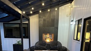 A wooden deck with a central table and chairs, a stone fireplace, and a covered area with string lights.