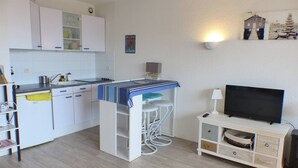 A compact kitchen with white cabinets, a built-in stove, and a bar area with stools. A flat-screen TV is on a white stand to the right. - CAPBRETON Residence "Les Terrasses de Notre-Dame", west facing with ocean views (CAPBRETON)