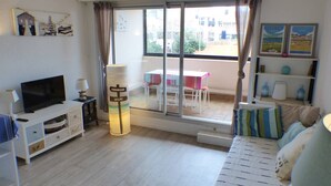 A living room with a TV, a lamp, a sofa, and a table with chairs. There is a window with curtains and a view of the outside. - CAPBRETON Residence "Les Terrasses de Notre-Dame", west facing with ocean views (CAPBRETON)