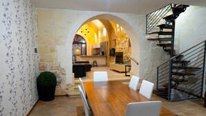 A dining area with a wooden table and white chairs, an arched doorway leading to a kitchen, and a staircase with metal railings. - Mixel Holiday Home 4 bedrooms with private pool (Ta' Sannat)