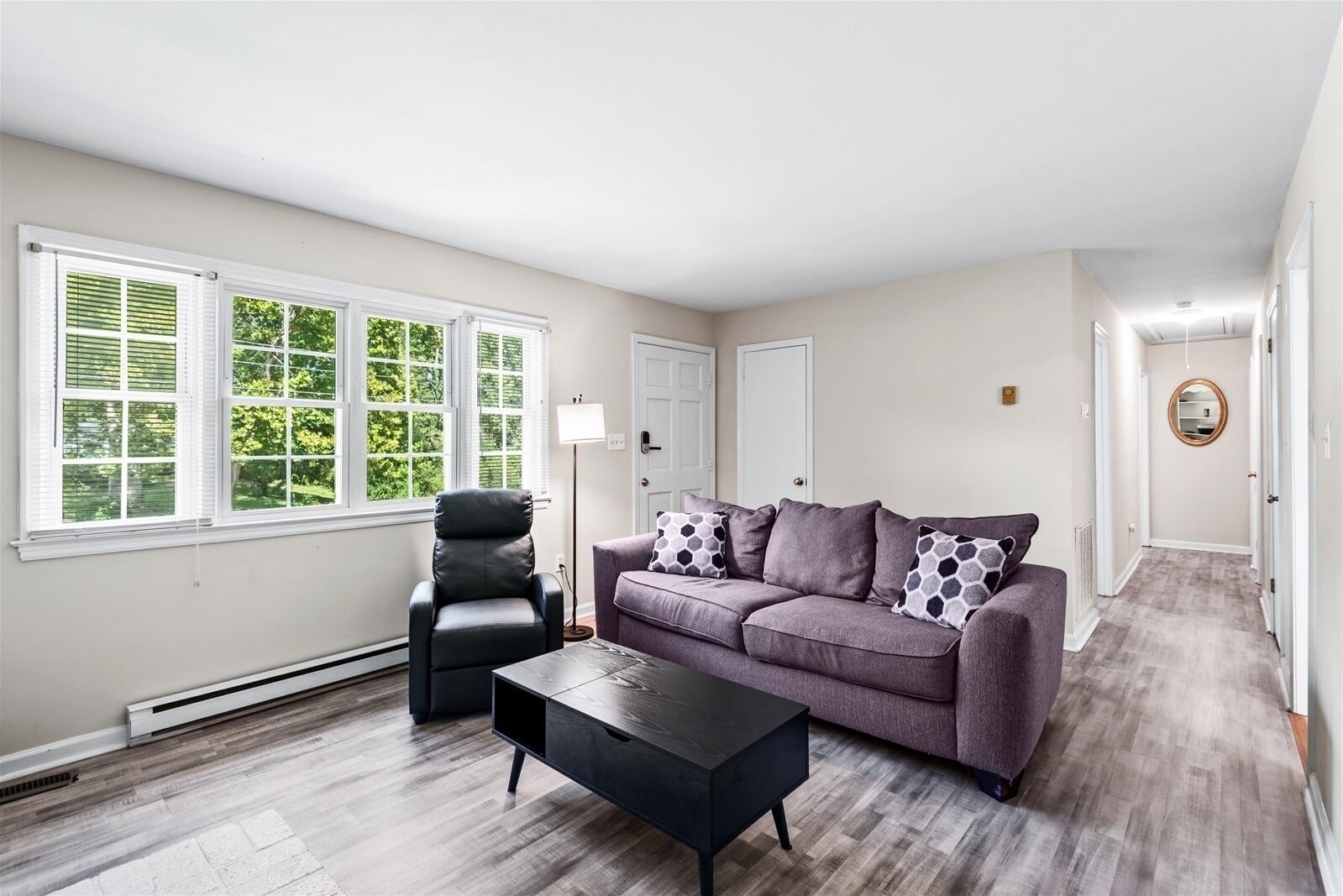 Un salon avec un canapé gris, un fauteuil noir et une table basse noire. De grandes fenêtres équipées de stores, une lampe sur pied et une porte sont visibles au fond.