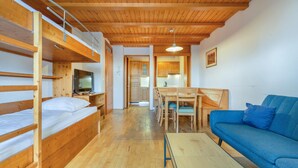 Quarto pequeno com beliches, televisão, mesa de jantar com cadeiras e um sofá azul.