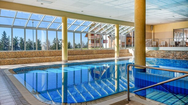 Piscina interior com paredes de vidro transparente, piso ladrilhado e vista para um edifício com varandas.