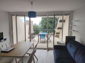 A modern living room with a sofa, a dining table with chairs, a TV, and a balcony with a table and chairs.
