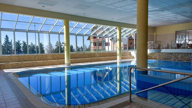 An indoor swimming pool with clear glass walls, a tiled floor, and a view of a building with balconies.