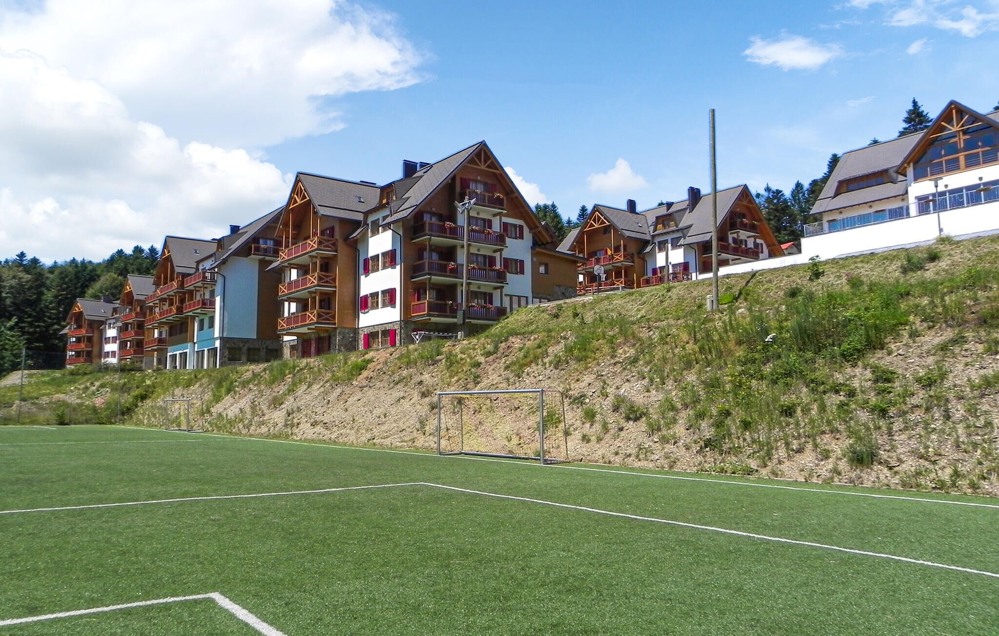 A row of multi-story buildings with balconies, situated on a hillside with a green field in the foreground.