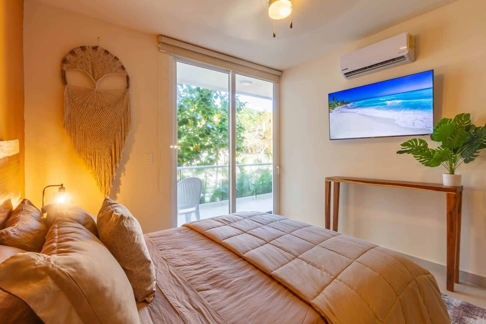 Un dormitorio con cama, ventana grande, aire acondicionado, televisor montado en la pared y planta en maceta.