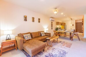A living room with a sofa, a coffee table, a dining table with chairs, and a kitchen area in the background.