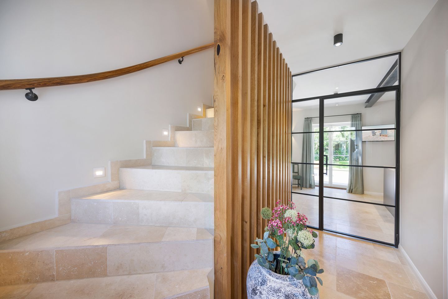 A modern staircase with beige tiles, a wooden handrail, and a potted plant with pink flowers.