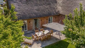 Una casa de ladrillo con techo de paja, un patio con mesa y sillas, y un jardín.