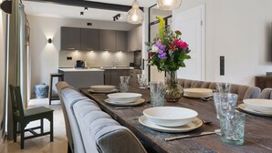 Un comedor moderno con una mesa larga dispuesta para seis personas, un jarrón de flores y una cocina al fondo.