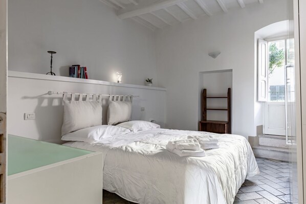A bedroom with a bed, a bedside table, a shelf with books, a lamp, and a window. - GuestHost - Bright apartment perfect for 4 people, with 2 bedrooms and 2 bathrooms, located in a period building in the heart of the historic center (ZTL). The apartment boasts all the main comforts, including a private garden, an oasis of peace and gree (Perugia)