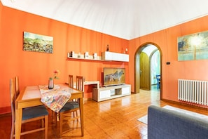 A living room with orange walls, a dining area with wooden furniture, a TV on a white stand, and a radiator. - GuestHost - Cozy and colorful 122 sqm apartment, capable of accommodating up to 6 people, located on the first floor of a building without an elevator. The location is enviable! We are just a few minutes' walk from Ursino Castle and Piazza Duomo. The nei (Catania)
