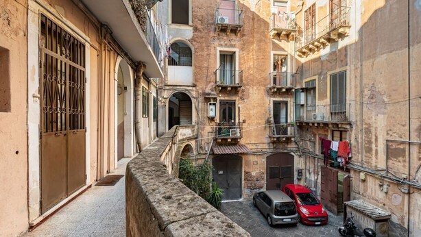 A narrow street with buildings on both sides, a stone wall on the left, and a small bridge. There are two cars parked on the right.