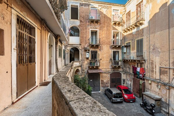 A narrow street with buildings on both sides, a stone wall on the left, and a small bridge. There are two cars parked on the right.