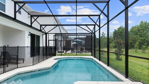 A swimming pool with a glass enclosure, a patio area with chairs, and a view of a house and greenery. - Stunning Modern Home! (Kissimmee)
