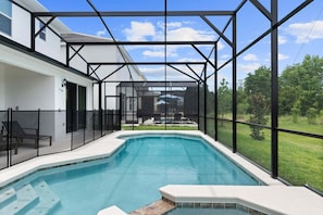 A swimming pool with a glass enclosure, a patio area with chairs, and a view of a house and greenery.