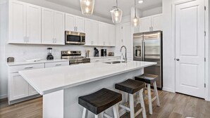 A modern kitchen with white cabinetry, stainless steel appliances, a large island with bar stools, and pendant lighting. - Lovely Townhome| Sleeps 8 (Davenport)