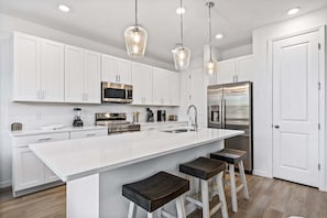 A modern kitchen with white cabinetry, stainless steel appliances, a large island with bar stools, and pendant lighting.