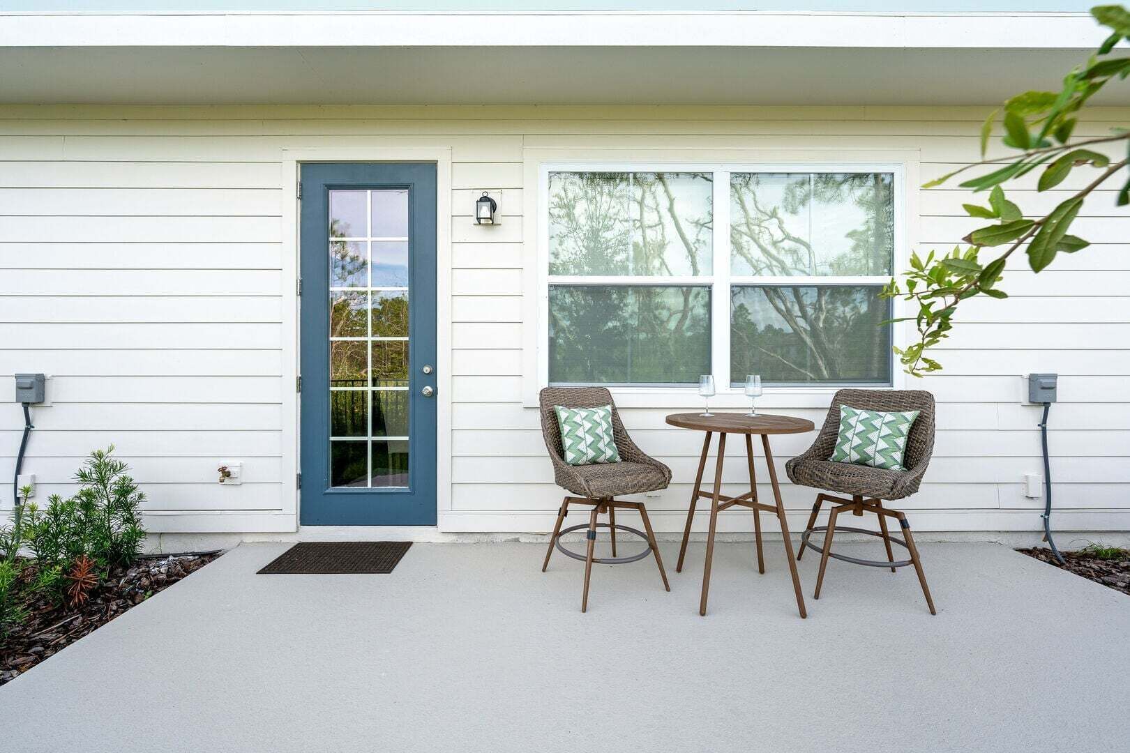 Una casa con puerta azul, dos sillones de mimbre con cojines, una mesita redonda y una ventana con vistas a árboles.