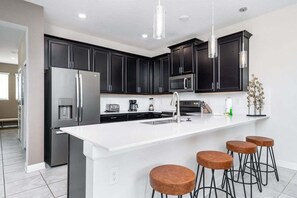A modern kitchen with dark cabinets, stainless steel appliances, a white island with stools, and pendant lighting.