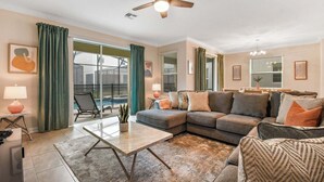 A living room with a sectional sofa, a coffee table, a floor lamp, a ceiling fan, and a view of a swimming pool.