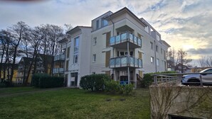 Un edificio residencial de varios pisos con balcones, un automóvil estacionado frente a él y un área verde con árboles.
