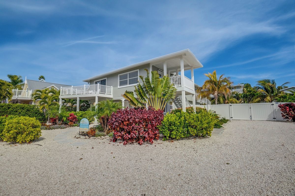 A two-story house with a balcony, surrounded by tropical vegetation and a gravel driveway.