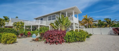 A two-story house with a balcony, surrounded by tropical vegetation and a gravel driveway.