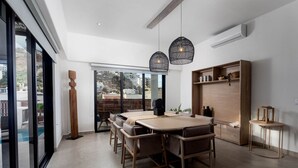 Un'area pranzo moderna con un grande tavolo di legno, diverse sedie, lampade a sospensione e vista sulla città attraverso ampie finestre.