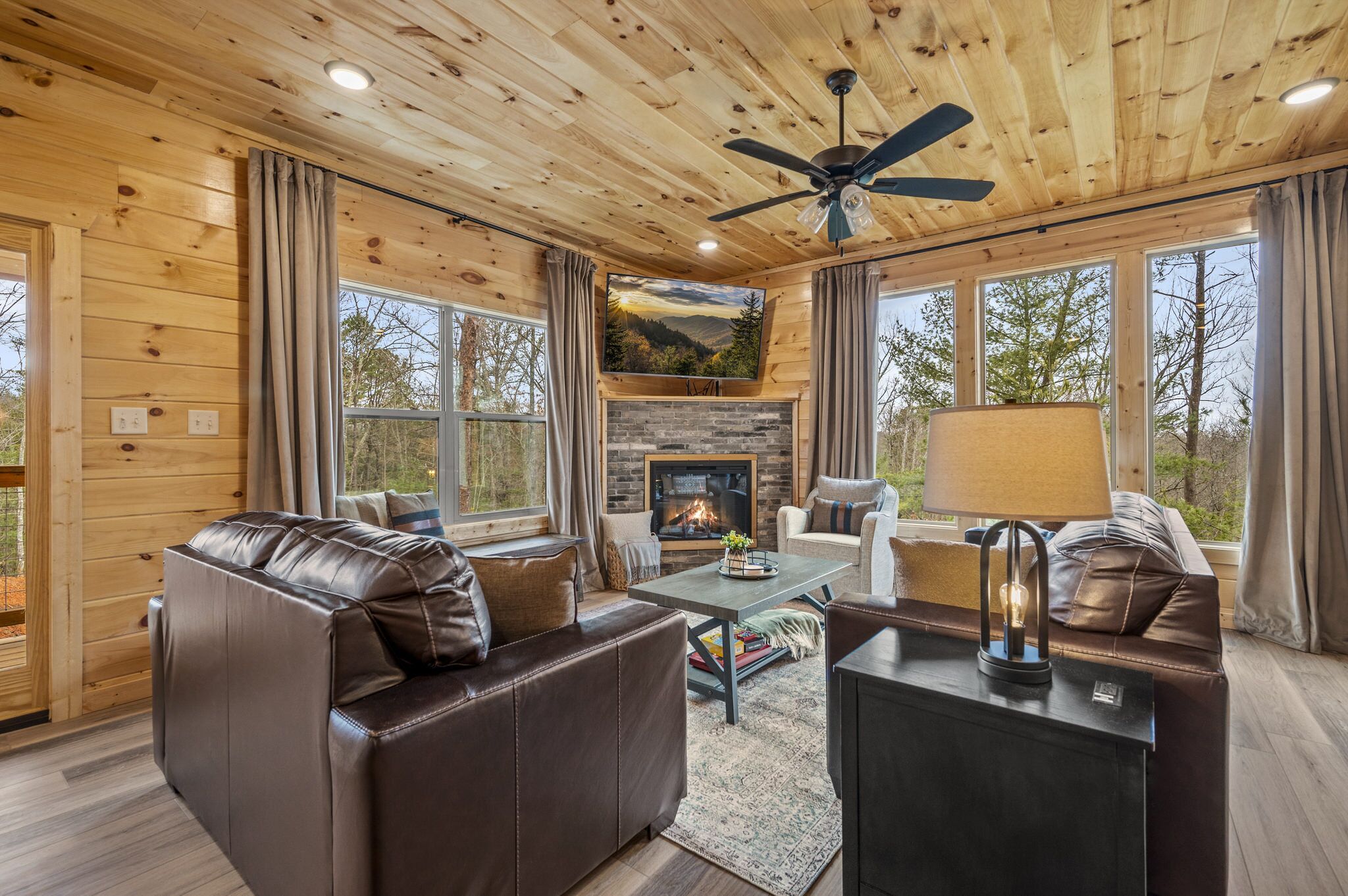 A cozy cabin interior with a fireplace, leather sofas, and large windows offering a view of the outdoors.