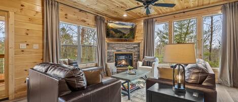 A cozy cabin interior with a fireplace, leather sofas, and large windows offering a view of the outdoors.